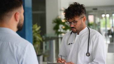 Tıbbi uzmanlar teşhisi tartışıyor, pano tutuyor, el kol hareketi yapıyor, modern hastane koridorunda duruyor, uzmanlık ve hasta bakımını iletiyor.