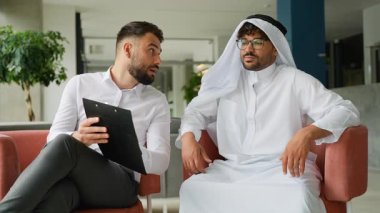 İki iş adamı, biri geleneksel Arap kıyafetleri içinde, diğeri beyaz gömlekli, modern bir ofis lobisinde rahat koltuklarda oturup, bir pano ile ilgili bir tartışmayla meşgul oluyorlar.