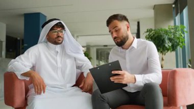 Biri geleneksel Arap kıyafetli, diğeri beyaz gömlekli iki işadamı, modern bir ofis lobisindeki rahat bir koltukta oturup, pano üzerindeki bir belgeyle ilgili bir tartışmaya giriştiler.