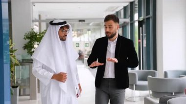 Biri geleneksel Arap kıyafetli, diğeri batılı iki işadamı, modern bir ofis binasında yürürken ciddi bir tartışma içindeler.