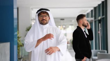 Geleneksel beyaz kandura giyen güvenilir emirati işadamı kollarını kavuşturup gülümserken batılı ortak telefonda modern ofis içi bulanık bir şekilde konuşuyor.