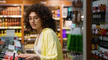Süpermarket alkol bölümünde şarap şişesini seçen genç bayan müşteri etiketi okuyor, karşılaştırıyor, satın alma konusunda karar veriyor, seçici odaklanıyor.