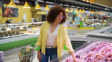 Kadın soğutulmuş market koridorunda paketlenmiş eti dikkatle inceliyor, bilgilendirilmiş ürün seçimi yaparken beslenme etiketini okuyor