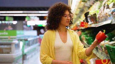 Bir süpermarkette kırmızı ve sarı dolmalık biber arasında seçim yapan genç bir kadın, karar vermeden önce onları dikkatle inceliyor.
