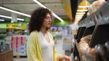 Genç bir kadın, marketin ekmek reyonunda dikkatlice ekmek seçerken, çoklu somun ve baget ekmeğine göz gezdirirken düşünceli bir satın alma kararı veriyor.