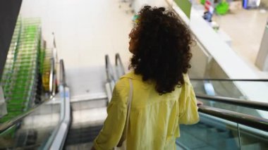 Kıvırcık saçlı ve gözlüklü kendinden emin bir kadın perakende sektöründe yürüyen yürüyen yürüyen merdiven, modern alışveriş alanında gezinirken neşeli bir gülümsemeyle geriye bakıyor.