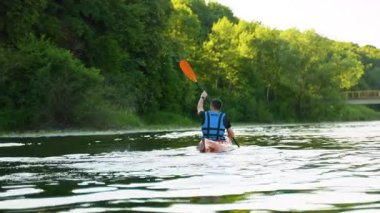 Koruyucu can yeleği giyen kanocu, yemyeşil orman manzarasıyla çevrili sakin nehir boyunca parlak turuncu bir kanoyla kürek çekiyor.