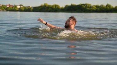 Sakallı adam gölde boğuluyor ve umutsuzca ayakta kalmaya çalışıyor, yardım istiyor ve nefes almak için nefes alıyor, suda nefes almaya çalışıyor.