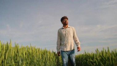 Kendine güvenen genç çiftçi, ekili yeşil buğday tarlalarında yürüyor ve etrafa bakıyor, çalışmalarının sonucunun keyfini çıkarıyor ve açık mavi gökyüzü altında gelecek ürünlerini planlıyordu.