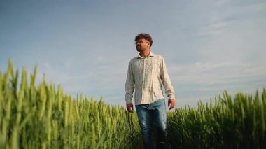 İyimser genç çiftçi gün batımında yeşil bir buğday tarlasında yürüyor ve gülümsüyor, çalışmalarının meyvelerinin ve doğanın güzelliğinin tadını çıkarıyor.