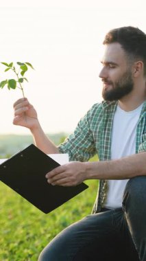 Tarım uzmanları bitki büyümesini inceliyor, tohum ve panoyu dikkatlice tutarken yeşil tarım arazisi çevresindeki gözlemleri belgeliyor.