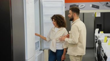 Modern buzdolabı modellerine göz atan genç çift, mutfak güncellemesi için ev aletlerini seçerken ilgili mağaza asistanından rehberlik alıyor.