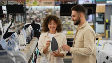 Gülümseyen çift ev aletleri bölümüne bakıyor, modern perakende elektronik ortamda yardımcı mağaza asistanı satın alma kararı ile yeni ütü seçiyor