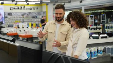 Evli çift televizyon modellerini dikkatlice değerlendiriyor, özellikleri ve özellikleri karşılaştırırken modern elektronik perakende ortamında birlikte alışveriş yapıyorlar.