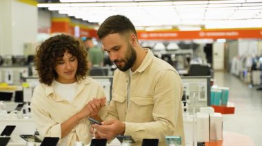 Modern teknoloji mağazasında akıllı telefon özelliklerini karşılaştıran genç bir çift, cihaz yeteneklerini keşfediyor ve büyük bir ilgiyle potansiyel alımı tartışıyorlar.