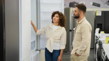 Modern eşyalar mağazasında buzdolabı alışverişi yapan çiftler, kadın özellikleri vurgularken, ortak ev mutfağı için ürün seçimini dikkatlice değerlendiriyor