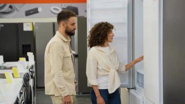 Mutlu müşteriler elektronik mağazasından yeni buzdolabı seçiyor, gülümseyen kadın buzdolabının kapısını açıp kapıyor, kocasıyla alışverişi tartışıyor, hipermarketteki ev aletlerini seçiyor.