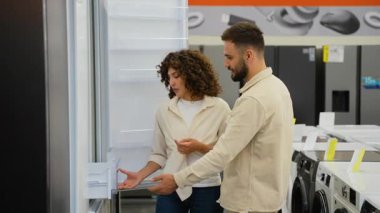 Genç bir aile eşyacı dükkanında yeni buzdolabı seçiyor, kadın buzdolabının kapısını açıyor ve kapanıyor, erkek karar vermede ona yardım ediyor.