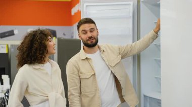Genç bir aile elektronik mağazasından yeni buzdolabı seçiyor, kadın raflarını gösteren ve bir şeyleri açıklayan bir adam, bir şeyi tartışıyor ve satın alma konusunda karar veriyorlar.