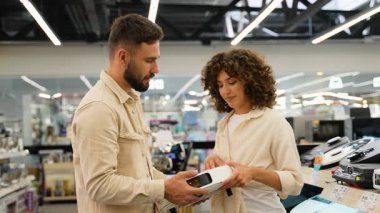 Gülümseyen çift elektronik mağazasında gelişmiş robot elektrik süpürgelerine göz atıyor, özelliklerini dikkatlice karşılaştırıyor ve kablosuz ev otomasyon teknolojisini tartışıyorlar.