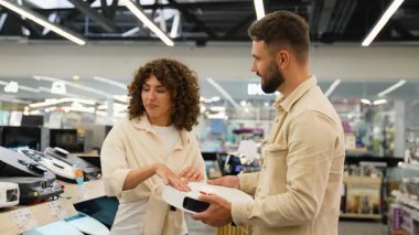 Gülümseyen çift elektronik mağazasında gelişmiş robot elektrik süpürgelerine göz atıyor, özelliklerini dikkatlice karşılaştırıyor ve kablosuz ev otomasyon teknolojisini tartışıyorlar.