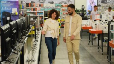 Genç bir aile bir elektronik mağazasından bilgisayar monitörü seçiyor, bir kadın bir erkeğe monitör gösteriyor, bir şeyi tartışıyorlar, bir erkek ve bir kadın mağazada geziniyorlar.