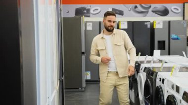 Sakallı adam, bej gömlekli adam, yeni buzdolabını seçiyor, buzdolabının kapısını açıp kapıyor, içine bakıyor, boyutunu ve özelliklerini değerlendiriyor.