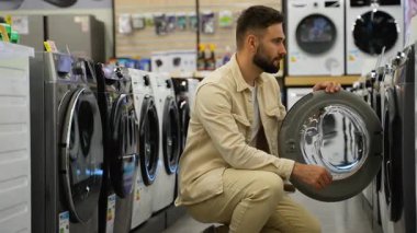 Genç adam bir elektronik mağazasında çeşitli çamaşır makinelerini dikkatlice inceliyor, kapıları açıyor ve kapanıyor, özelliklerini kontrol ediyor ve satın almadan önce modelleri karşılaştırıyor.
