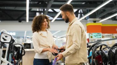 Müşteriler elektrik süpürgesi özelliklerini ve performanslarını dikkatlice karşılaştırırken elektronik mağaza görüntülerini inceleyip olası ev temizlik çözümlerini değerlendiriyorlar.