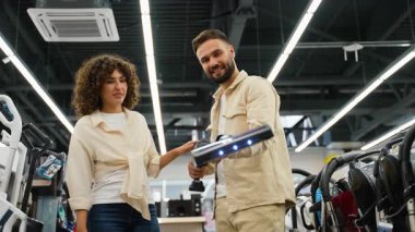 Mutlu çift elektronik mağazasında yeni kablosuz elektrik süpürgesini test ediyor, gülümsüyor ve özelliklerini tartışıyor, ev aletlerinin alımı hakkında karar veriyor.
