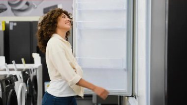 Tatmin olmuş bayan müşteri modern ev aletleri perakende galerisinde mutfak teknolojisi seçimi yaparken şık buzdolabı modelini dikkatlice inceliyor.