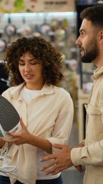 Mutlu müşteriler bir mağazadan yeni bir ütü seçiyorlar, bir tezgahtar doğru seçimi yapmalarına yardım ediyor, ütüyü ellerinde tutuyorlar ve dikkatlice bakıyorlar.