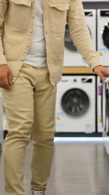 Günlük giysiler içinde genç sakallı bir adam çömelmiş çamaşır makinelerini inceliyor, bir mağazada davul çalıyor, ev aletlerini seçme sürecini gösteriyor.