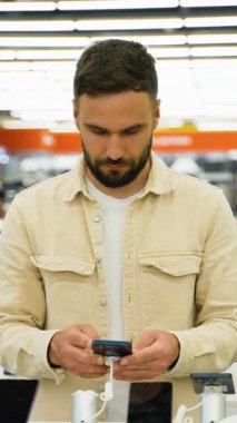Günlük bej ceketli ve beyaz tişörtlü sakallı adam, parlak ışıklandırılmış bir elektronik mağazasında sergilenen çeşitli akıllı telefonları inceliyor.