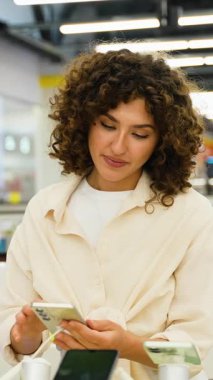 Elektronik mağazasında yeni bırakılmış akıllı telefonu test eden kadın, cihaz ekranını dikkatle inceleyen ve yenilikçi mobil teknoloji özelliklerini keşfeden kadın