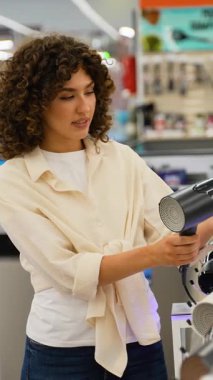 Genç bir kadın elektronik mağazası vitrinine göz atarken şık saç kurutma makinesini dikkatlice değerlendiriyor, alışveriş deneyimi sırasında ürün özelliklerini dikkatle analiz ediyor.