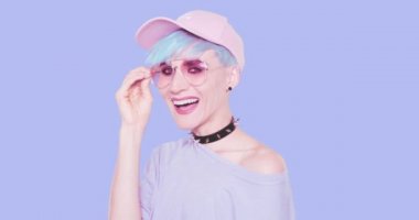 Moda alternatifi sırıtan mavi saçlı, şık güneş gözlüklü ve şapkalı genç kız stüdyoda mavi arka planda poz veriyor. Şehir tarzı görünüş ve aksesuar konsepti