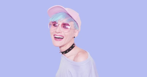 Moda alternativa sonriente chica feliz con el pelo azul, gafas de sol con estilo y la tapa posando en el estudio sobre fondo azul. Aspecto elegante urbano y concepto de accesorios — Vídeo de stock