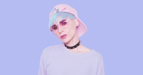 Fashion Swag Teenager verspieltes Mädchen mit blauen Haaren, stylischer Sonnenbrille und Mütze posiert im Studio vor blauem Hintergrund. Urban stylischer Look und Accessoire-Konzept — Stockvideo