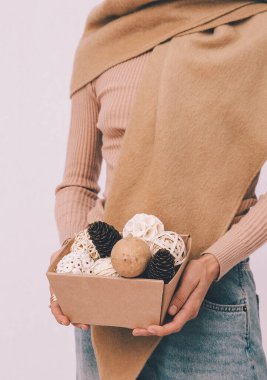 Bej rengi sıcak süveter ve atkı giyen genç bir kız, beyaz uzayda topları olan bir Noel süsü kutusu tutuyor. Minimum moda festivali Noel / Yeni Yıl konsepti.