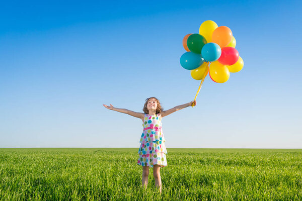 Happy child playing with bright multicolor balloons outdoor. Kid having fun in green spring field against blue sky background. Healthy and active lifestyle concept