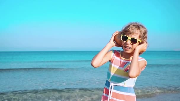 幸せな子供は ビーチで貝殻を聞きます 青い海と空の背景の女の子の肖像画 子供の夏休みに楽しい時を過します 夢と想像力の概念 — ストック動画