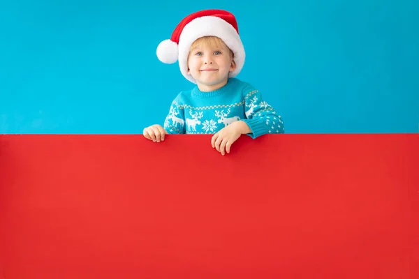 Niño feliz sosteniendo banner rojo de Navidad en blanco contra espalda azul — Foto de Stock