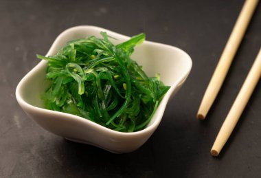 Seçici odak noktası, beyaz seramik tabaklarda susam tohumlu deniz Chuka salatası, koyu arkaplanda bambu çubukları.