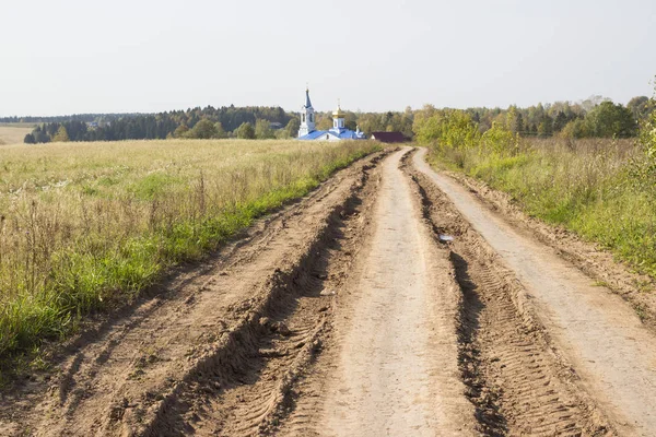 Road on a grass covered land with church steeple and rooftop background. Russian field.