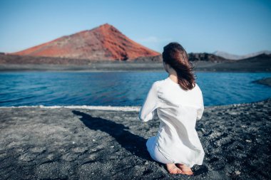 Back view of woman in white clothes praying on black sand beach Playa Bermeja, Lanzarote, Canary Islands, Spain clipart