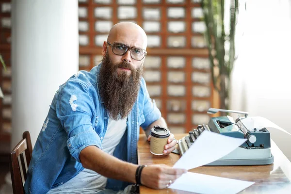 Bearded stylish writer with cup of coffe reading his novel. Modern writer in glasses working on new book in library