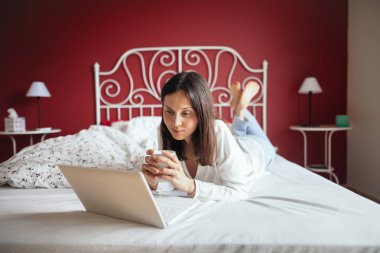 Yatağa rahatlatıcı ve içme kahve dizüstü bilgisayar ile evde düşünceli kadın. Geri kalan kavramı