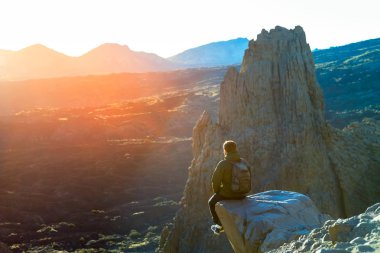 Genç seyyahı uçurumun kenarında oturan adam ve dağlarda günbatımı keyfini çıkarın. Seyahat kavramı