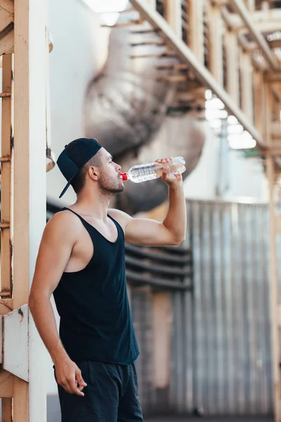 年轻人从瓶子里喝水 在工业城市背景下锻炼锻炼后的运动员休息 — 图库照片